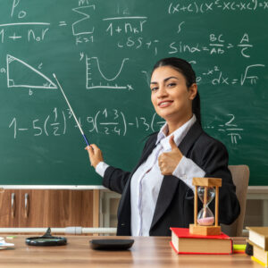 smiling young female teacher sits table with school supplies points blackboard with pointer stick showing thumb up classroom